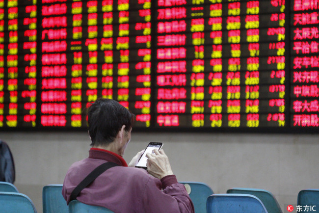 A Chinese investor looks at his smartphone in front of an electronic display showing prices of shares (red for price rising and green for price falling) at a stock brokerage house in Nanjing city, east China's Jiangsu province, 17 October 2018. [Photo: IC]