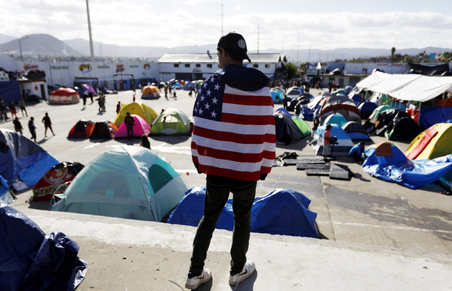 A man from Honduras wears a U.S. flag around his shoulders as he looks out over migrant tents at a former concert venue serving as a shelter in Tijuana, Mexico, Dec. 2, 2018. [Photo: AP/Rebecca Blackwell]