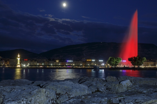 The famous water fountain Le Jet d'Eau is illuminated in the color red on the occasion of the 70th anniversary of the Geneva Conventions with the moon seen in the background, in Geneva, Switzerland, 12 August 2019. [Photo: EPA via IC/SALVATORE DI NOLFI]